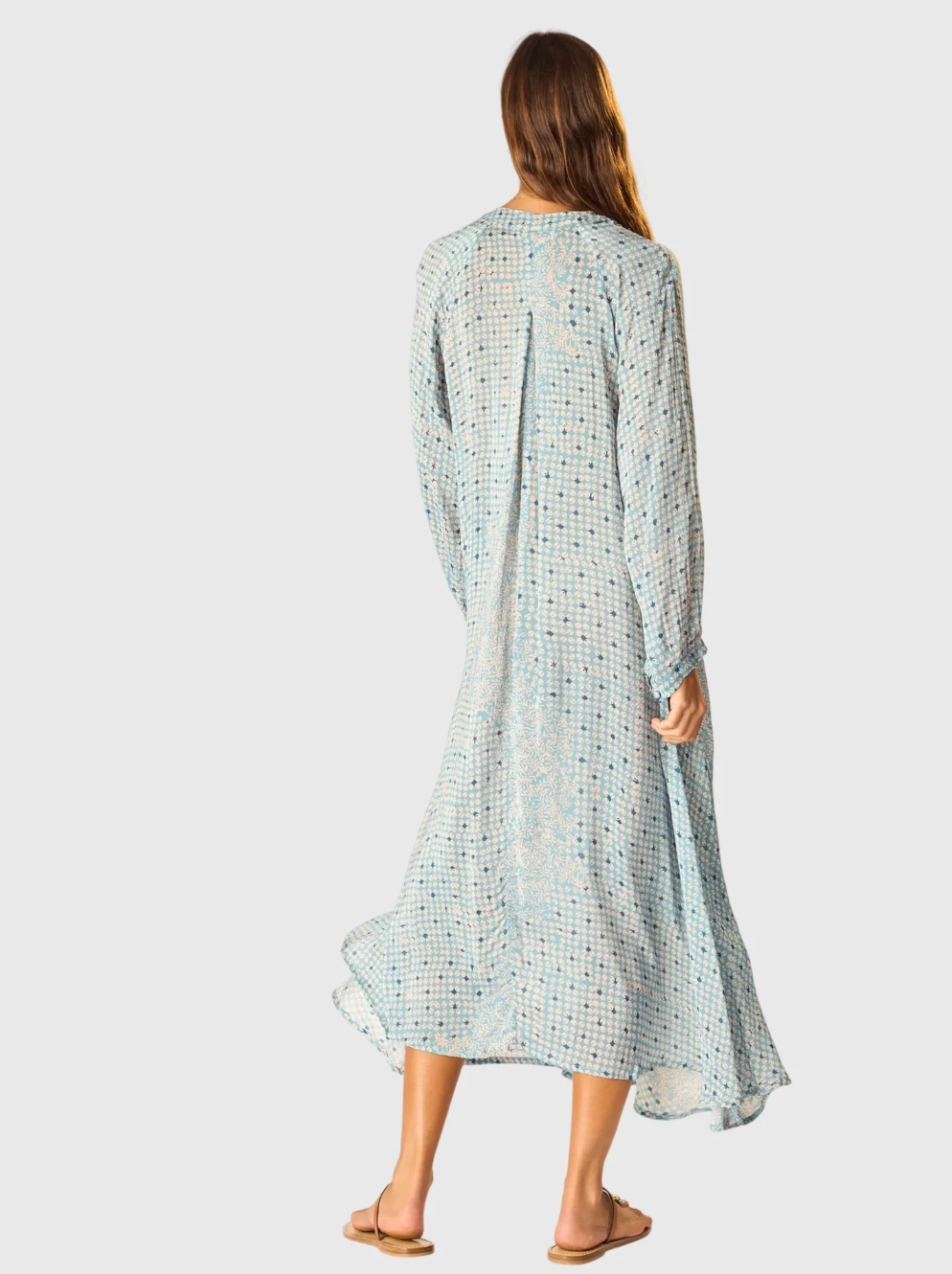 A woman with long hair stands with her back to the camera in a natalie martin Fiore Maxi—a loose, light blue patterned A-line dress with a v-neckline and long sleeves—paired with sandals against a plain background.