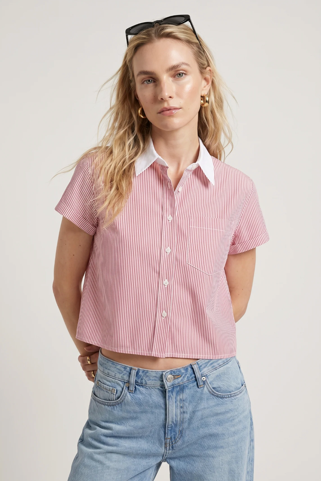 A woman with long blonde hair and sunglasses models THE SHIRT’s Boxy Short Sleeve Cropped Shirt—red and white striped, featuring No Gape button technology—paired with light blue jeans against a plain background.