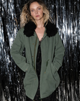 Oversized olive green Le Superbe Utility Anorak with black faux-fur collar, worn over a black dress.