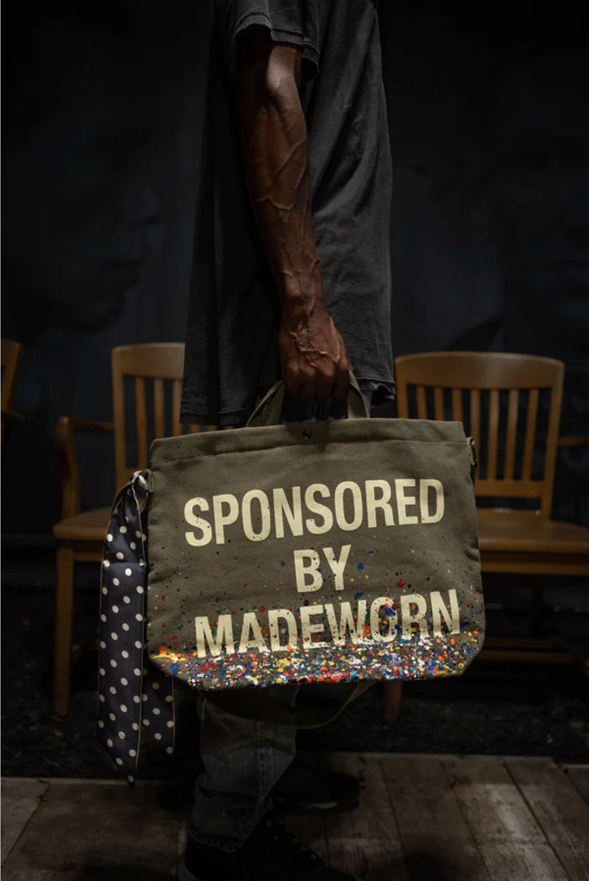 A person holds the Made Worn Artist Tote, a limited-edition olive-green cotton canvas bag with bold white "SPONSORED BY MADEWORN" text and colorful paint splatters; wooden chairs sit in the dim background.