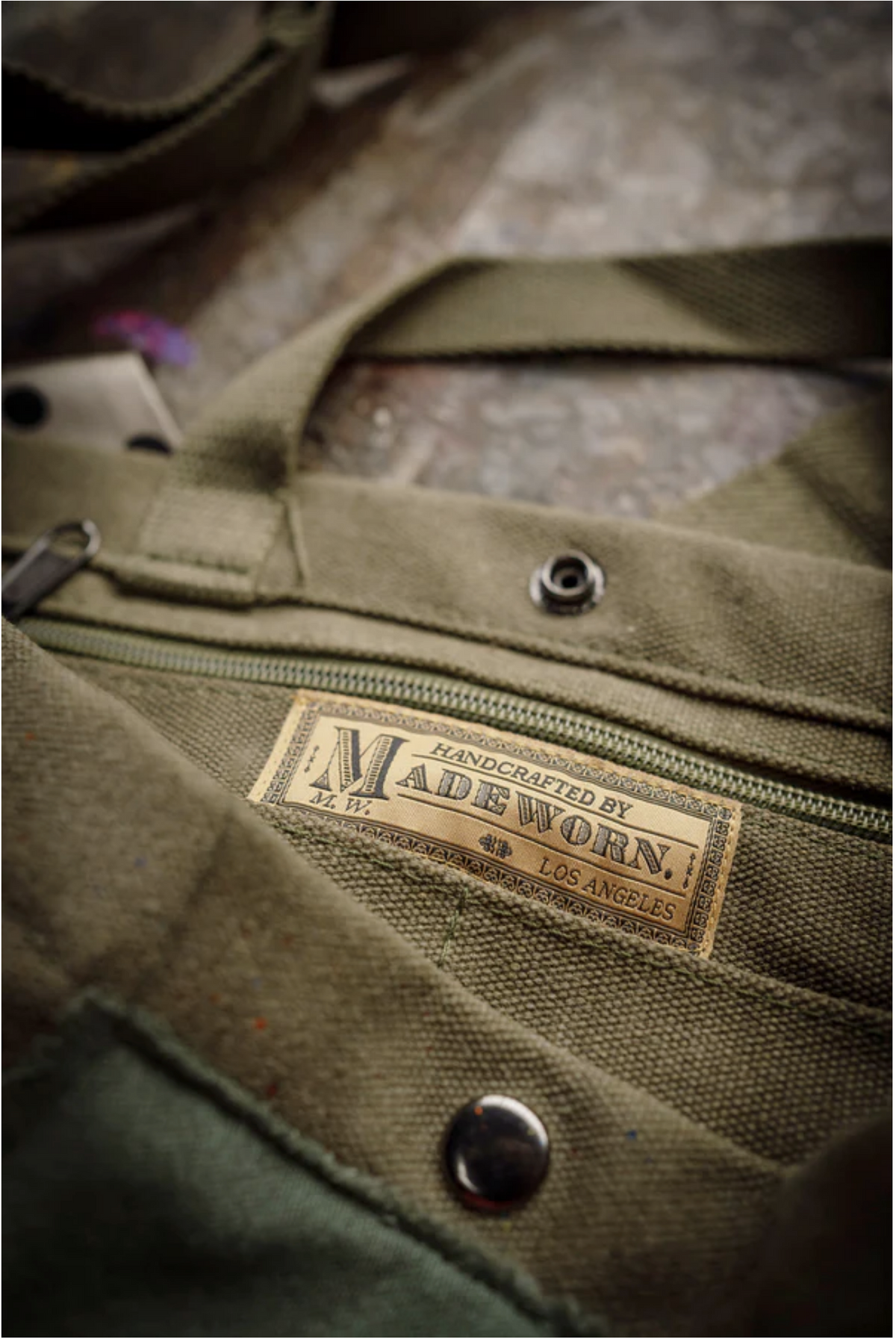 A close-up of the limited-edition Artist Tote by Made Worn shows a label reading "Handcrafted by MadeWorn, Los Angeles" stitched near the zippered pocket and metal snap button, emphasizing its craftsmanship.