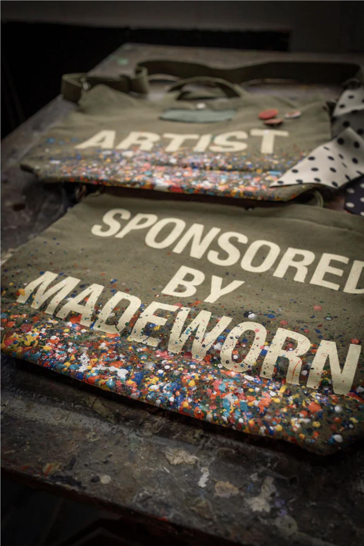 Two limited edition Artist Tote bags by Made Worn, crafted in olive-green cotton canvas with colorful paint splatters—one reads “ARTIST,” the other “SPONSORED BY MADEWORN”—are displayed on a paint-splattered surface.