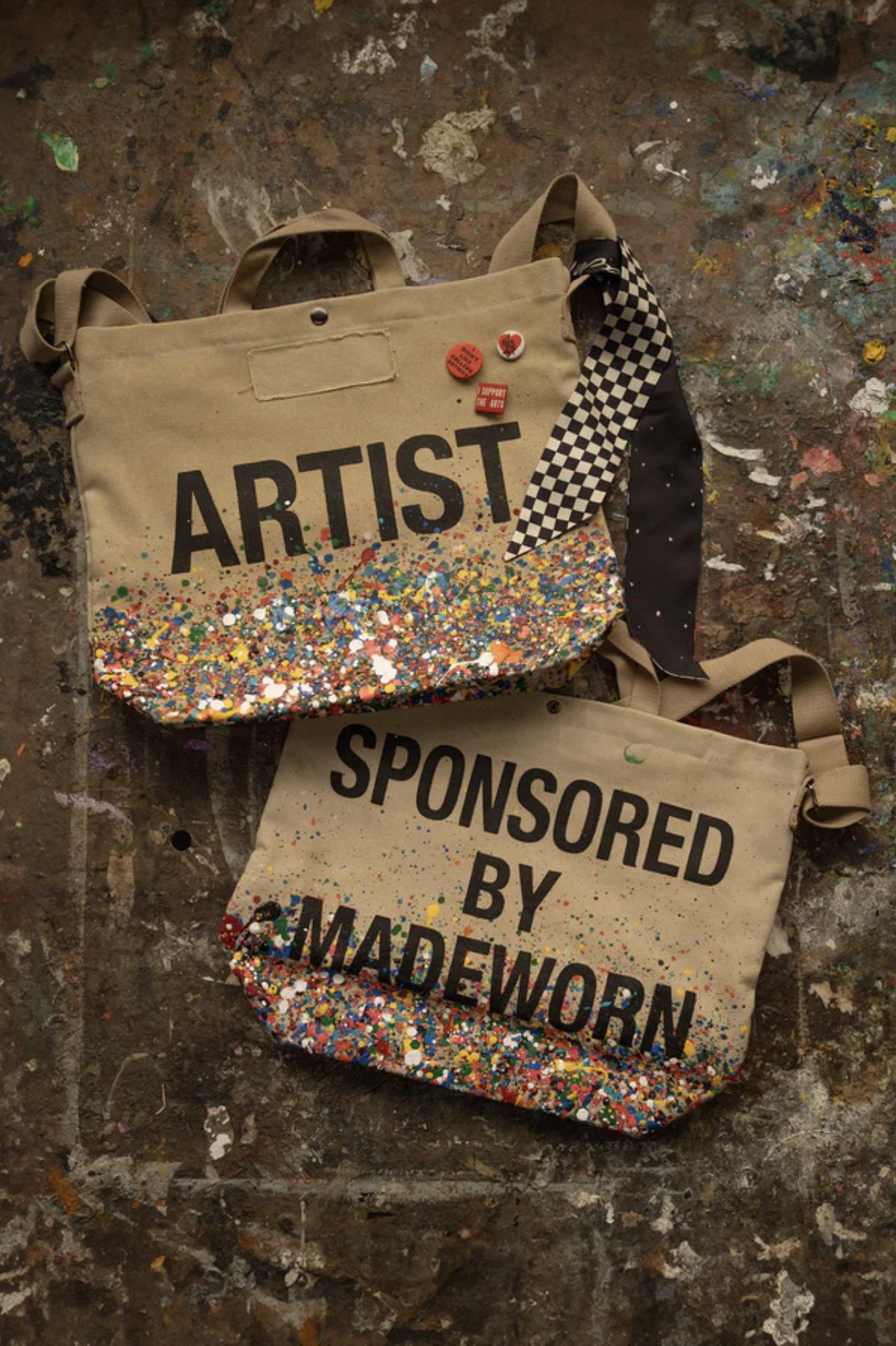 Two beige cotton canvas Artist Tote bags by Made Worn sit on a paint-splattered surface. One reads "ARTIST" in colorful glitter, the other "SPONSORED BY MADEWORN," both sparkling with similar decorative accents.