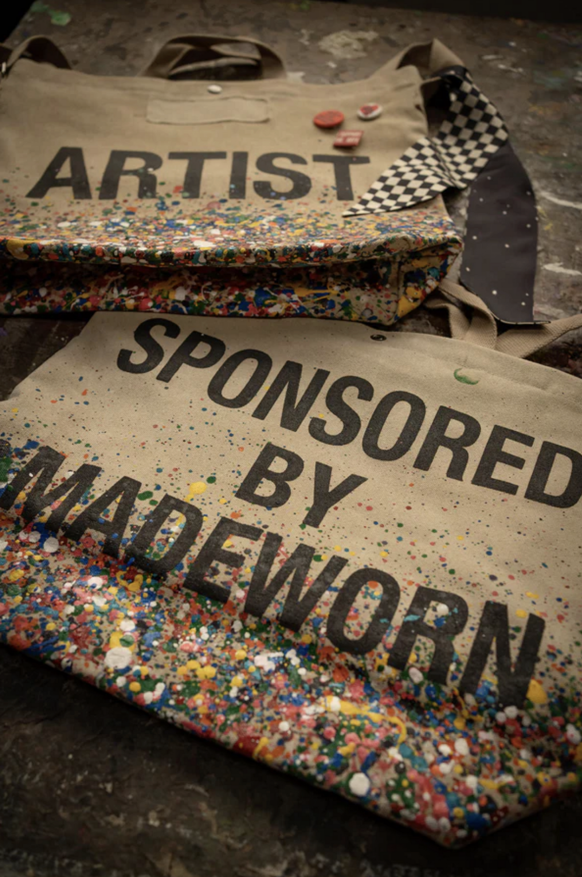 Two limited-edition beige cotton Artist Tote bags by Made Worn with colorful paint splatters—one features "ARTIST," the other "SPONSORED BY MADEWORN." The totes are displayed on a textured surface with custom buttons and a checkered cloth.