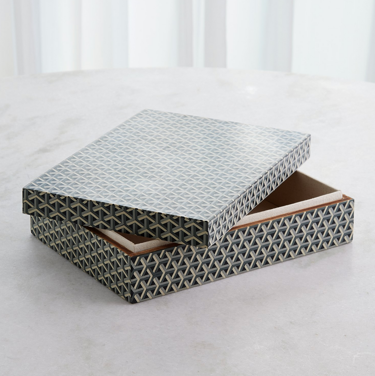 The Global Views Interwoven Box LG, featuring a geometric blue and white design, rests on a light surface with its lid partially open to show the empty interior.