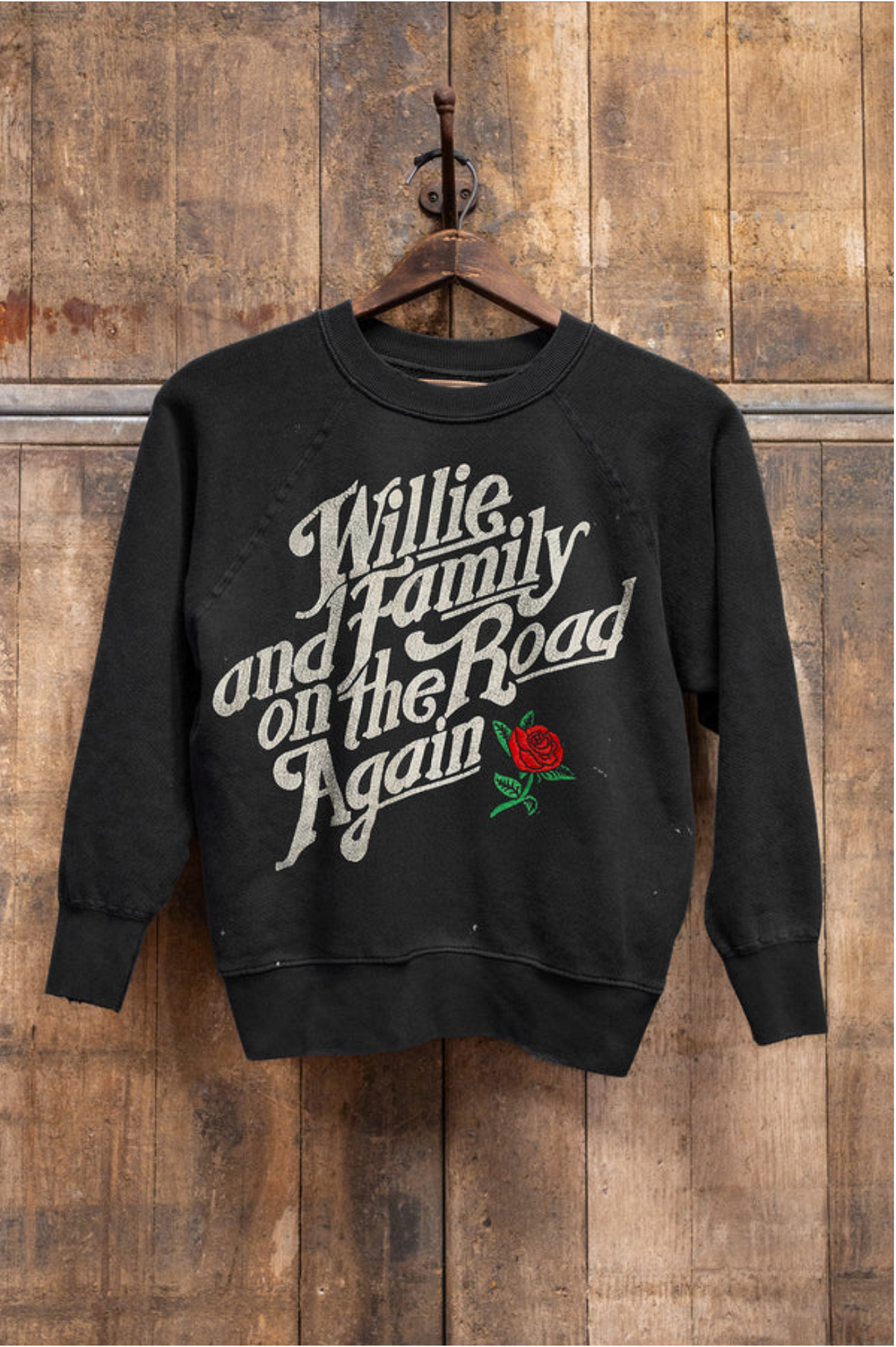 The Madeworn Shrunken Sweatshirt by Made Worn, crafted from heavyweight cotton, showcases "Willie and Family on the Road Again" in white retro font with a red rose graphic, hanging on a wooden hanger against a rustic wooden wall.