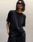A person with wet, chin-length dark hair wears black sunglasses, black pants, and Sold Out NYC's The Oversized Tee—known for its buttery-soft feel—standing by a light stone wall with one hand in their pocket.