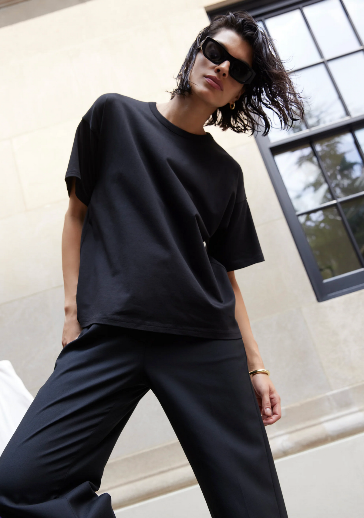 A person with short, wavy hair wearing black sunglasses, black pants, and The Oversized Tee by Sold Out NYC stands outdoors in front of a light stone building with a large window, leaning slightly forward.