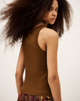 A young woman with voluminous curly hair captures modern boho style in the sleeveless brown BA&SH Top Cynda and a colorful patterned skirt, standing sideways and glancing over her shoulder against a plain backdrop.