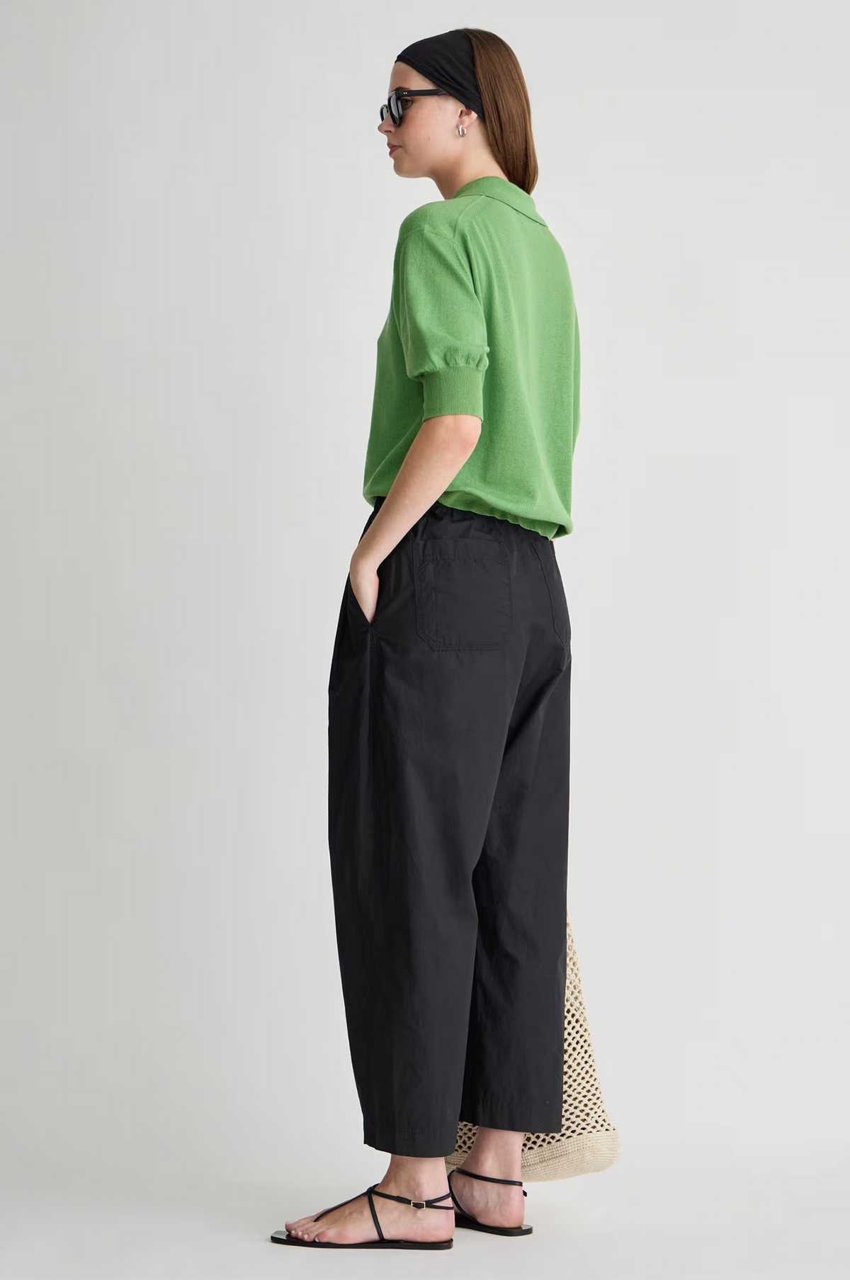 A woman stands sideways with hands in pockets, wearing Apiece Apart’s Spa Pleat Pant with an elastic waistband and a green polo shirt. She accessorizes with black sandals, sunglasses, a black headband, and a woven beige bag against a plain light background.