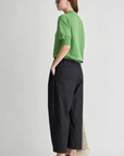 A woman stands sideways with hands in pockets, wearing Apiece Apart’s Spa Pleat Pant with an elastic waistband and a green polo shirt. She accessorizes with black sandals, sunglasses, a black headband, and a woven beige bag against a plain light background.