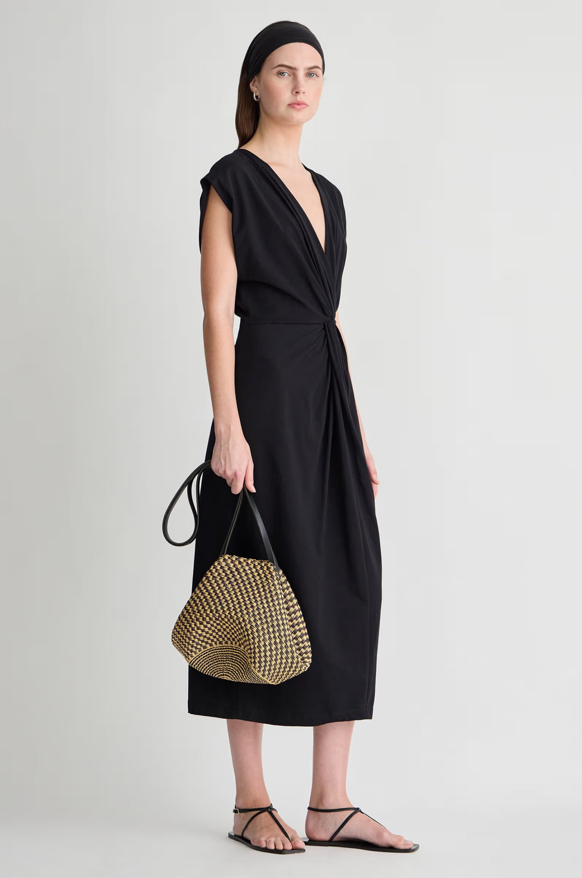 A woman in the Apiece Apart Margit Twist Dress, paired with a black headband, black sandals, and a woven straw handbag with black handles, stands against a plain light background.