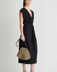 A woman in the Apiece Apart Margit Twist Dress, paired with a black headband, black sandals, and a woven straw handbag with black handles, stands against a plain light background.