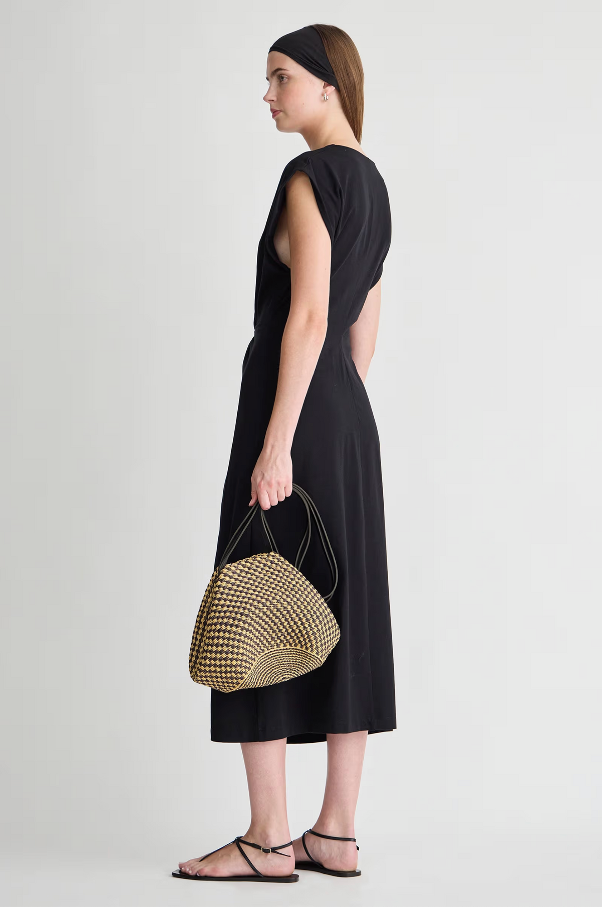 A woman stands sideways against a plain light background wearing the Apiece Apart Margit Twist Dress in black cotton jersey, paired with black sandals, a black headband, and holding a woven dome-shaped handbag.