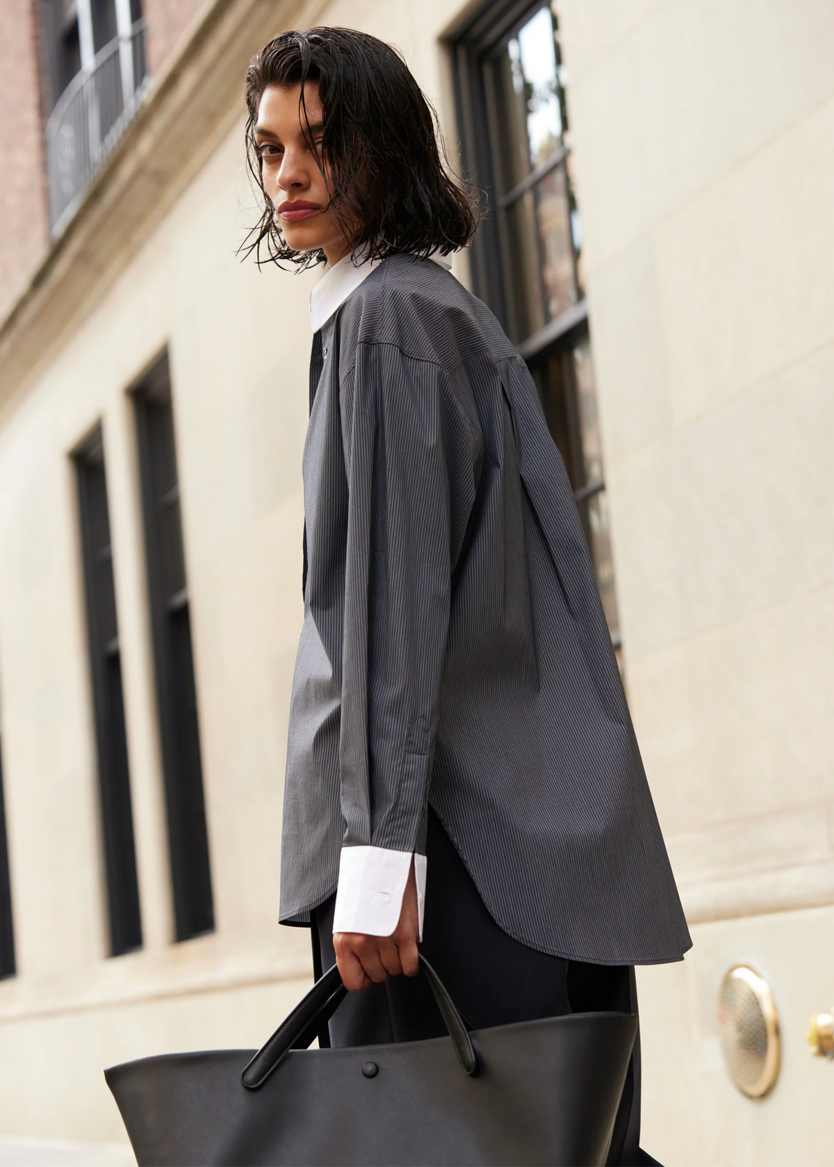 A person with wet, dark hair stands outside by a stone building, wearing the oversized Sold Out NYC "The Everything Shirt" in gray pinstripes with white cuffs and collar, and carrying a large black tote bag.
