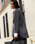 A person with wet, dark hair stands outside by a stone building, wearing the oversized Sold Out NYC "The Everything Shirt" in gray pinstripes with white cuffs and collar, and carrying a large black tote bag.