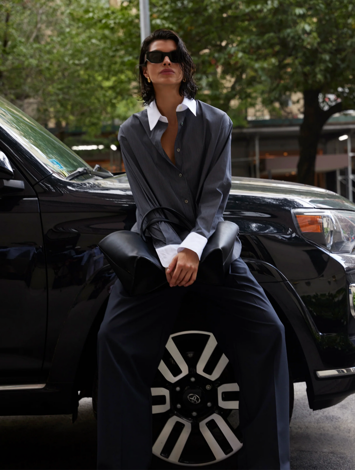 Someone wearing The Everything Shirt by Sold Out NYC and sunglasses sits on the hood of a black SUV, holding a large black bag with green trees and city buildings in the background.