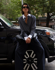Someone wearing The Everything Shirt by Sold Out NYC and sunglasses sits on the hood of a black SUV, holding a large black bag with green trees and city buildings in the background.