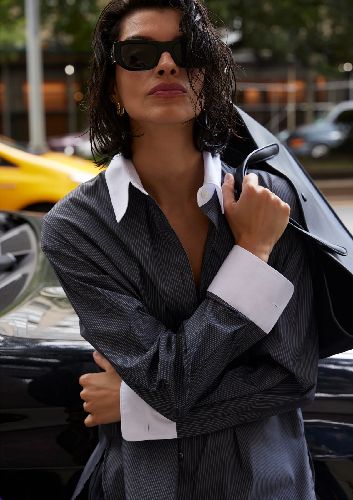 A woman with wet, chin-length hair and dark sunglasses wears The Everything Shirt by Sold Out NYC—an oversized pinstripe style with crisp white cuffs and collar—carrying a jacket with a blurred yellow car in the background.