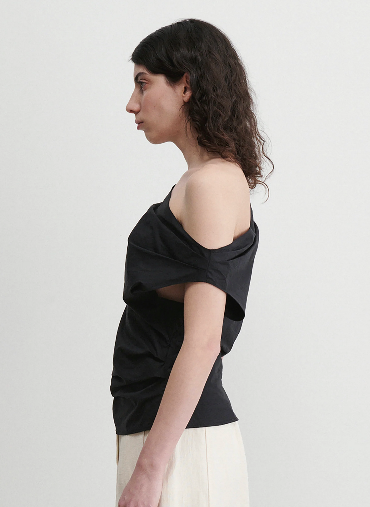 A person with shoulder-length curly hair stands in profile against a plain background, wearing the Rachel Comey Mata Top and light-colored pants.