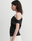 A person with shoulder-length curly hair stands in profile against a plain background, wearing the Rachel Comey Mata Top and light-colored pants.
