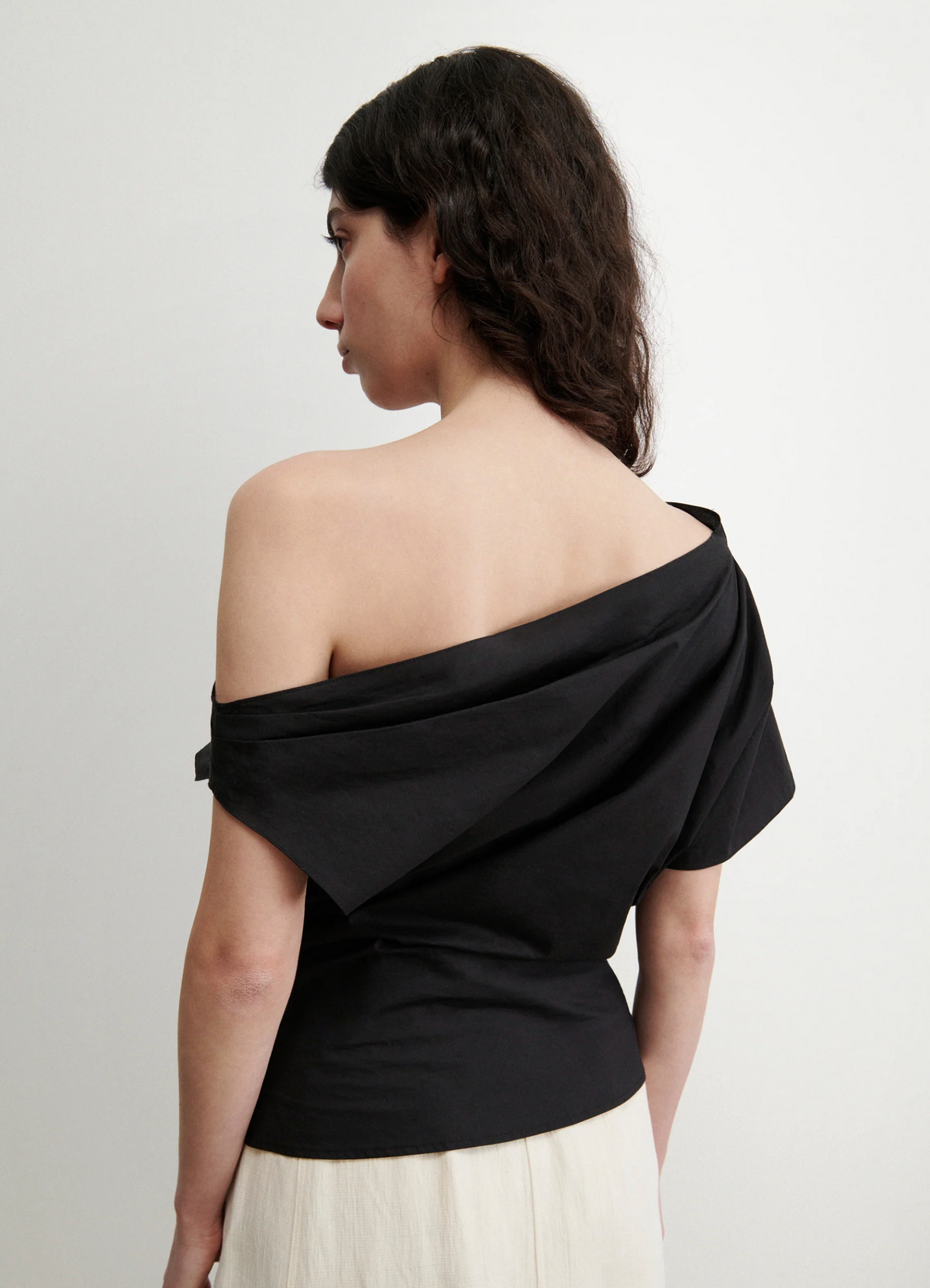 A woman with wavy dark hair faces away, wearing the black Mata Top by Rachel Comey and a light organic cotton skirt, standing against a plain white background.