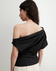 A woman with wavy dark hair faces away, wearing the black Mata Top by Rachel Comey and a light organic cotton skirt, standing against a plain white background.