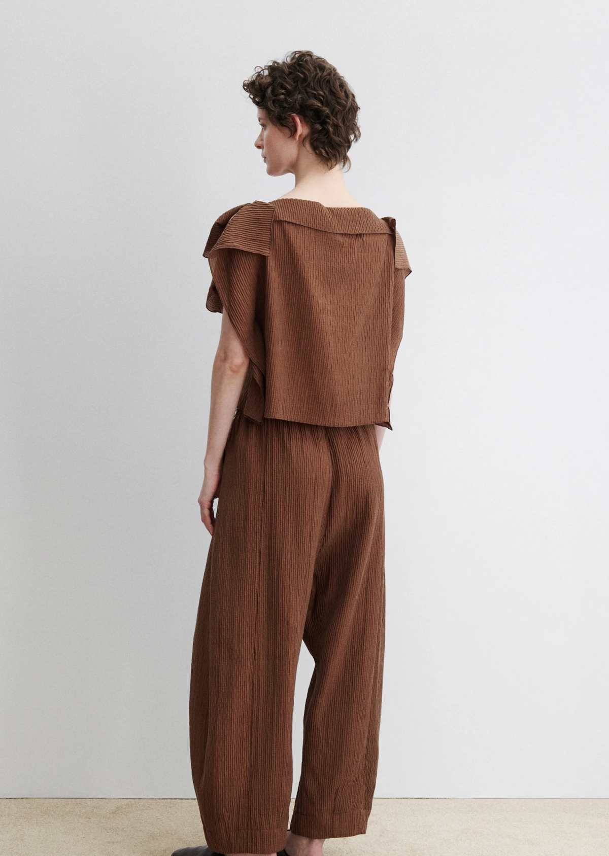 A person with short curly hair stands sideways, wearing the Rachel Comey Rivera Top—a relaxed crinkled blouse with wide flutter sleeves—paired with matching pants, against a plain light background.