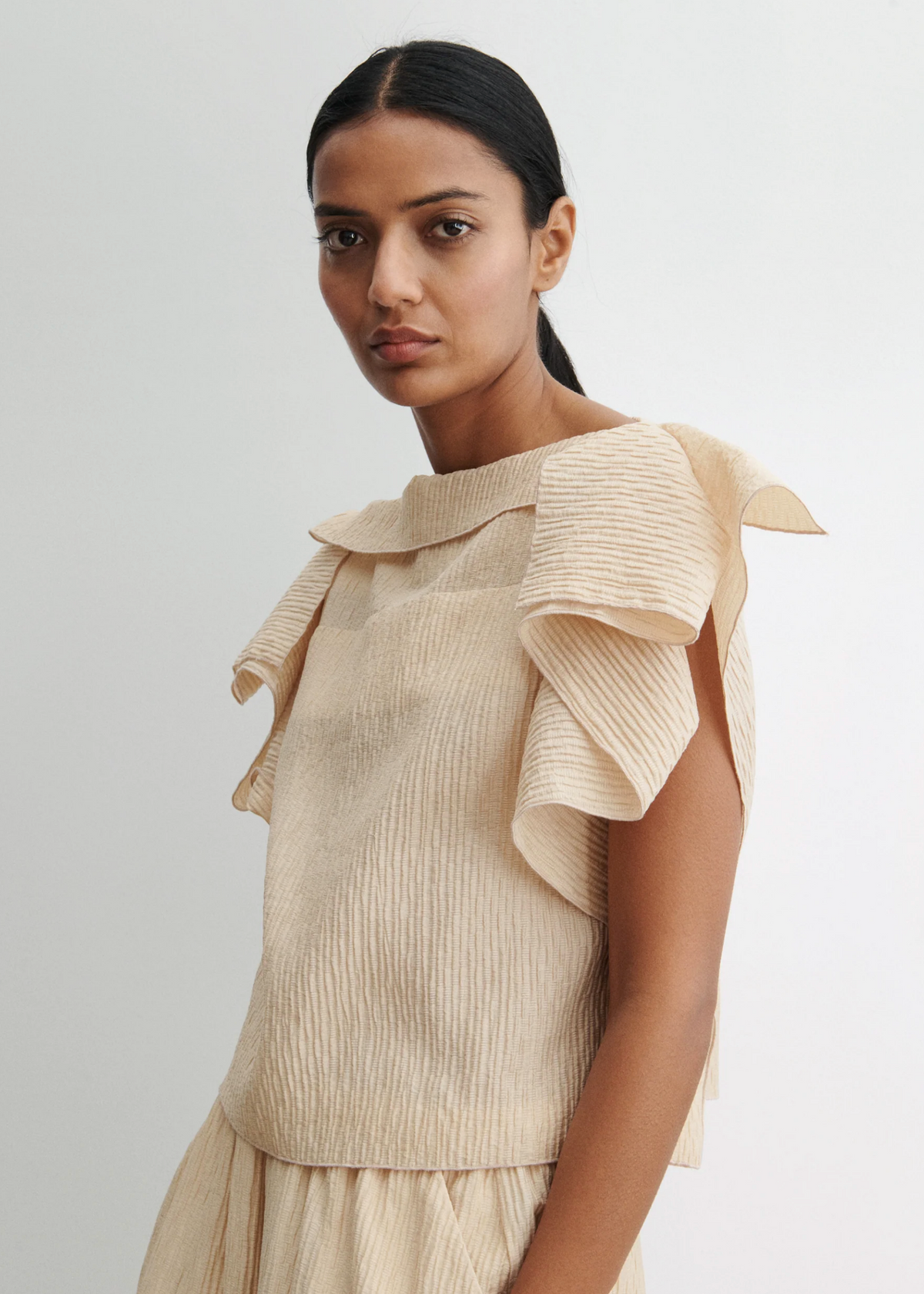 A woman with long dark hair tied back wears the Rachel Comey Rivera Top, an airy beige blouse in crinkled fabric with large ruffled flutter sleeves. She stands against a plain light background, looking calmly at the camera.