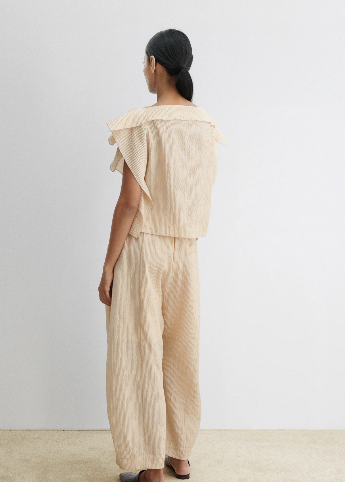 A person with long dark hair tied back is seen from behind, wearing the Rachel Comey Rivera Top in beige with layered flutter sleeves and matching wide-leg pants, against a white wall and beige floor.