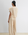 A person with long dark hair tied back is seen from behind, wearing the Rachel Comey Rivera Top in beige with layered flutter sleeves and matching wide-leg pants, against a white wall and beige floor.