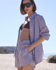 A woman stands on a sandy beach in Sold Out NYC’s The Everything Shirt, an oversized blue-and-white striped cotton button-down, paired with matching shorts and sunglasses; hills and blue sky form the backdrop.