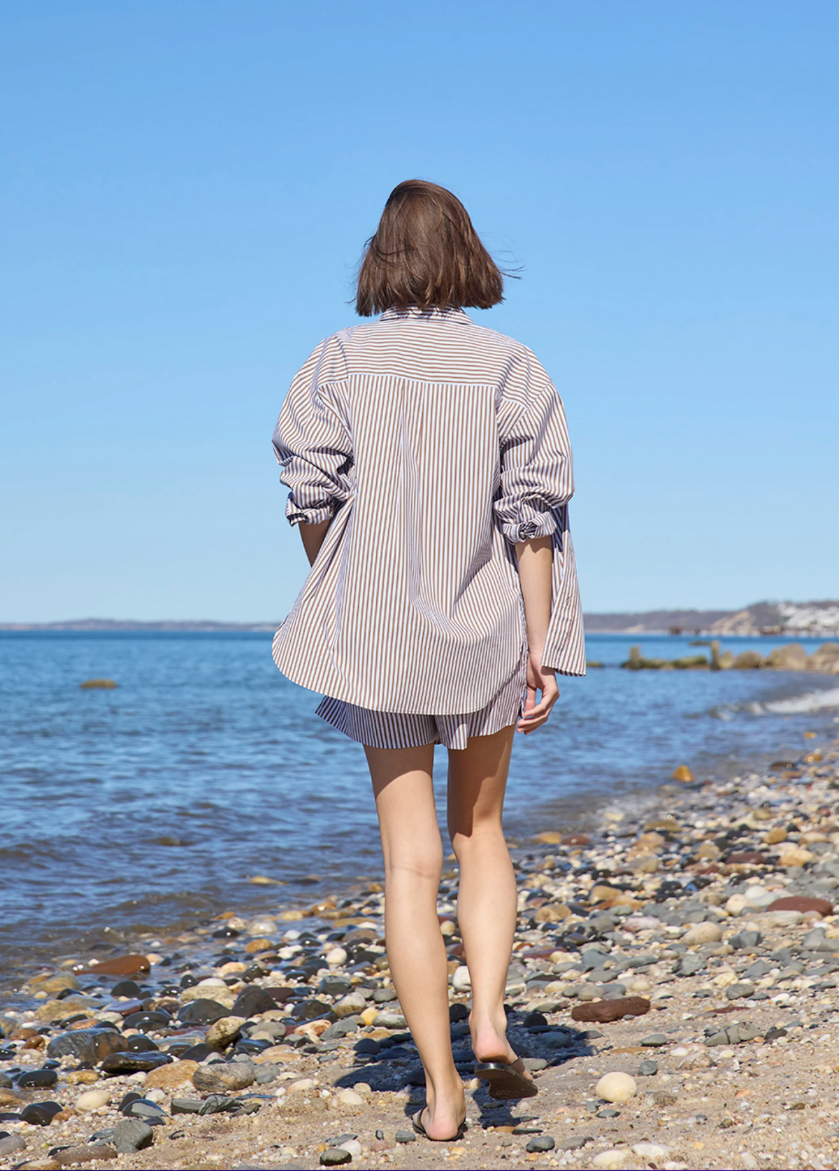 Wearing Sold Out NYC's The Everything Shirt, a person with short brown hair strolls barefoot across a rocky beach toward the ocean beneath a clear blue sky.