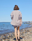 Wearing Sold Out NYC's The Everything Shirt, a person with short brown hair strolls barefoot across a rocky beach toward the ocean beneath a clear blue sky.