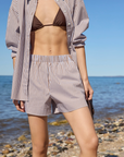 A person stands by the shore in The Everything Shirt by Sold Out NYC, styled over a brown bikini top and matching striped shorts. The calm sea and rocky beach stretch out beneath a clear blue sky.