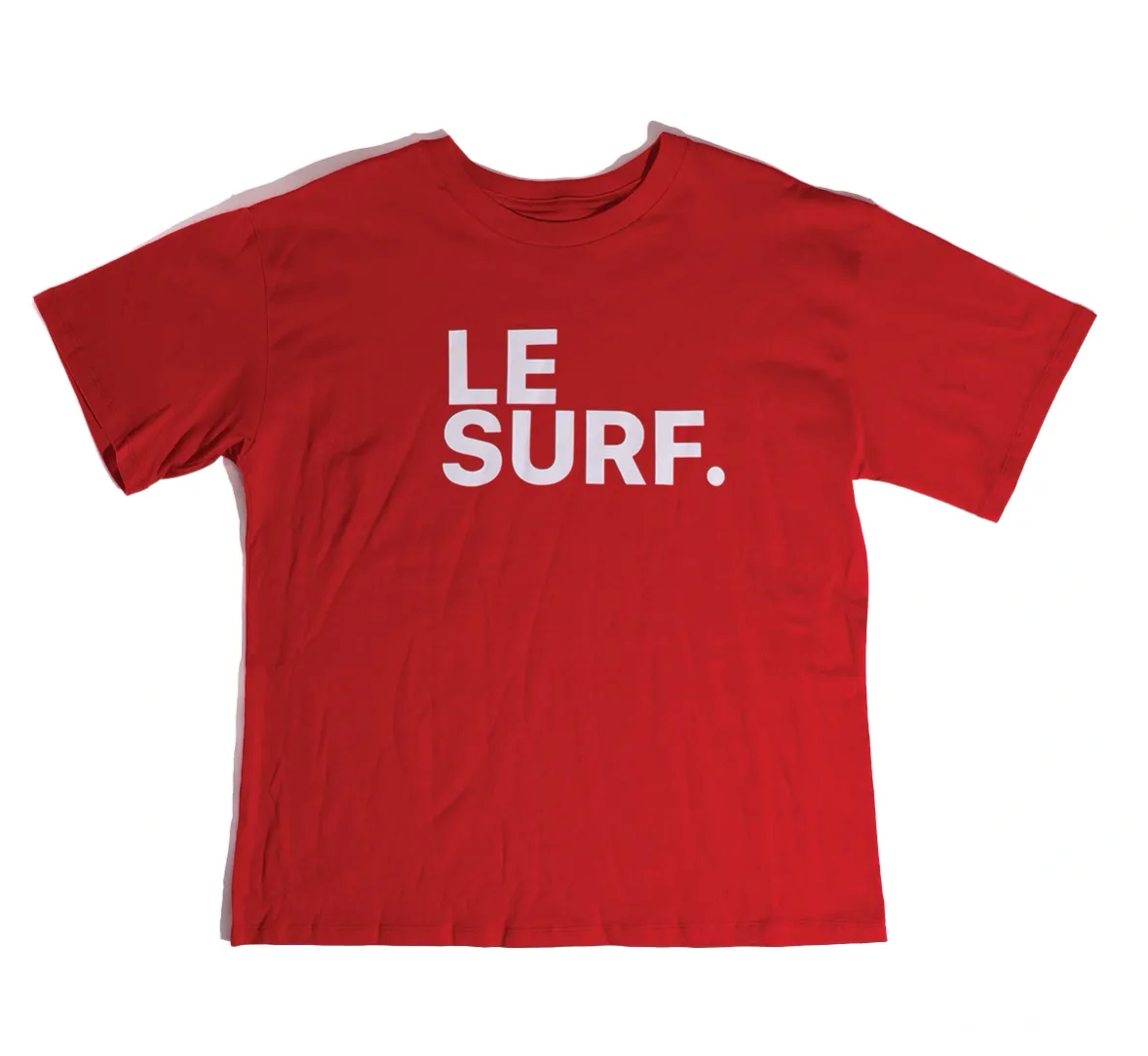 The Le Surf Tee by Ping Pong Surf Club is a loose-fit red T-shirt made from soft combed cotton, featuring "LE SURF." in bold white capital letters across the chest.