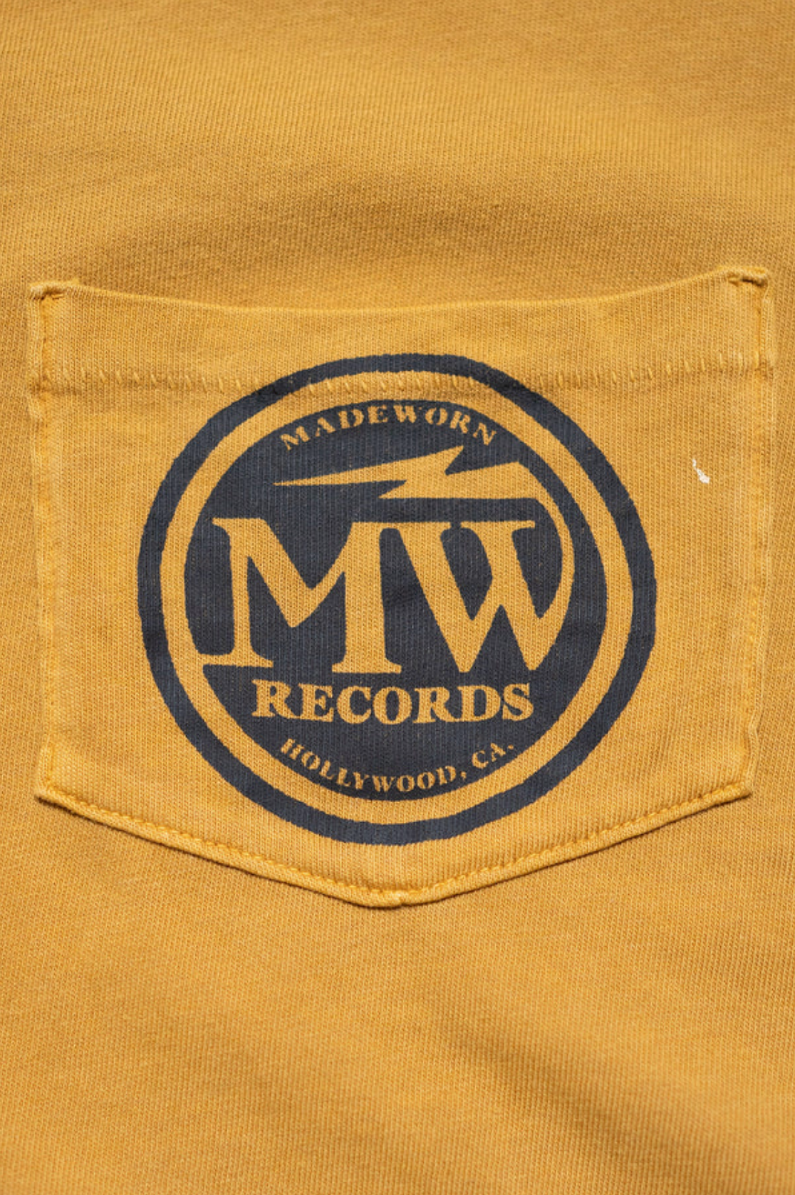 The Made Worn Pocket Tee MadeWorn Records is a 100% cotton, unisex vintage-style tee made in the USA, featuring a mustard yellow denim pocket with a blue circular "MADEWORN MW RECORDS HOLLYWOOD, CA" logo.