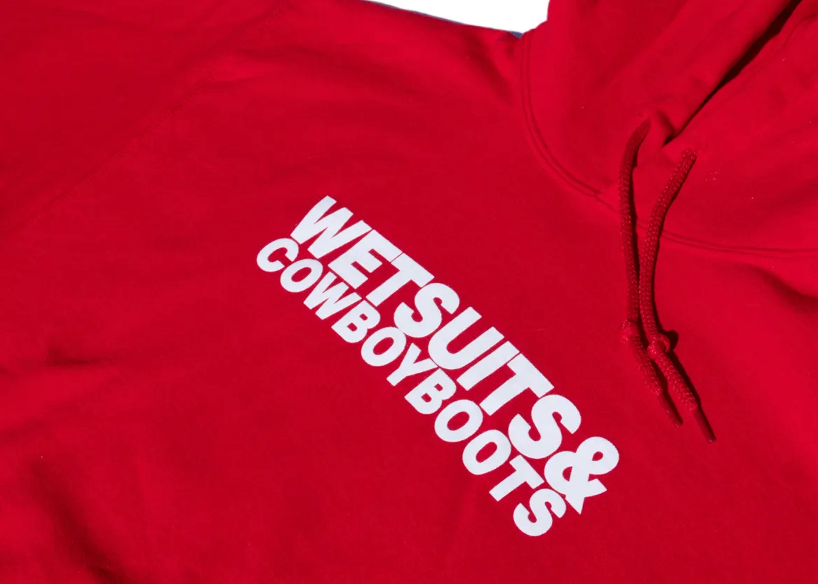 The Wet Suit & Cowboy Boots Hoodie by Ping Pong Surf Club is a red, iconic piece with "WETSUITS & COWBOY BOOTS" in bold white lettering, made from a soft cotton-polyester blend.