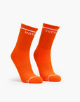 A pair of bright orange "Baby Steps" socks by Mother, with white ribbed cuffs and bold "MOTH" on the left and "FUCK" on the right sock in capital letters near the top.