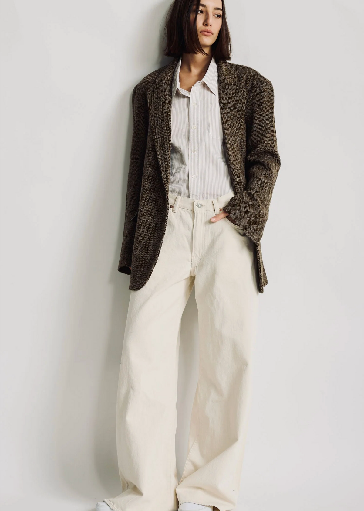 A person with straight dark hair wears the Denimist HYANNIS DECONSTRUCTED BLAZER over a striped shirt and loose, high-waisted cream pants, standing against a plain white background.