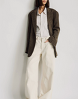 A person with straight dark hair wears the Denimist HYANNIS DECONSTRUCTED BLAZER over a striped shirt and loose, high-waisted cream pants, standing against a plain white background.