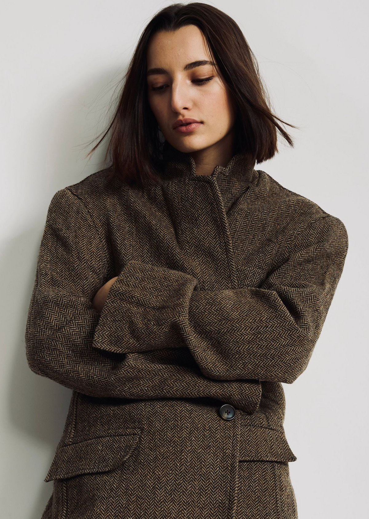 A person with straight, shoulder-length dark hair stands against a light background, wearing Denimist’s HYANNIS DECONSTRUCTED BLAZER—an oversized herringbone style with a classic lapel collar—their arms crossed and head tilted slightly downward.