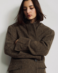 A person with straight, shoulder-length dark hair stands against a light background, wearing Denimist’s HYANNIS DECONSTRUCTED BLAZER—an oversized herringbone style with a classic lapel collar—their arms crossed and head tilted slightly downward.