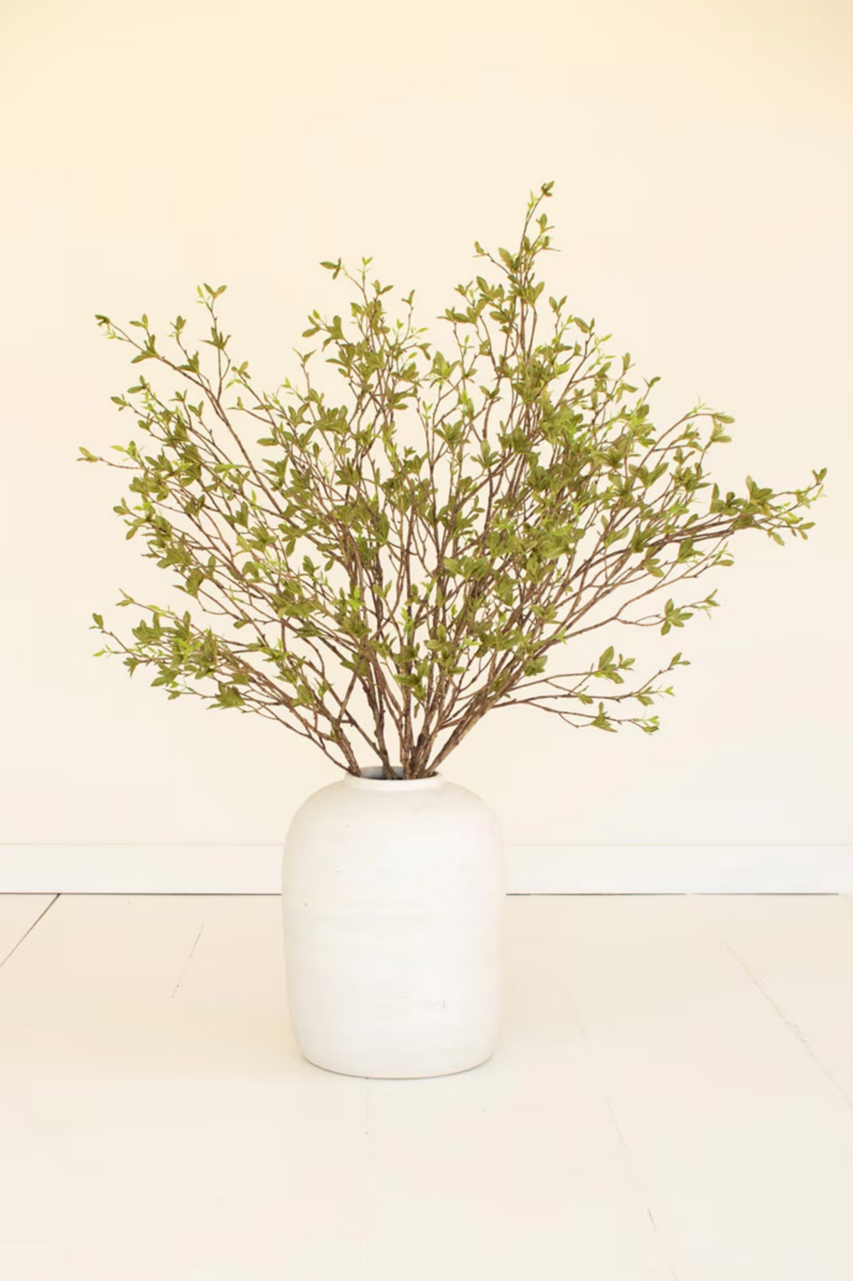 White vase with Kalalou Budding Leaf Spray on a white floor, set against a neutral beige background.
