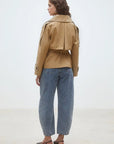 A woman with wavy brown hair models the SUNCOO Elliott Jacket, a belted short trench coat in tan, paired with blue cuffed jeans and black heeled shoes, standing against a plain light background.