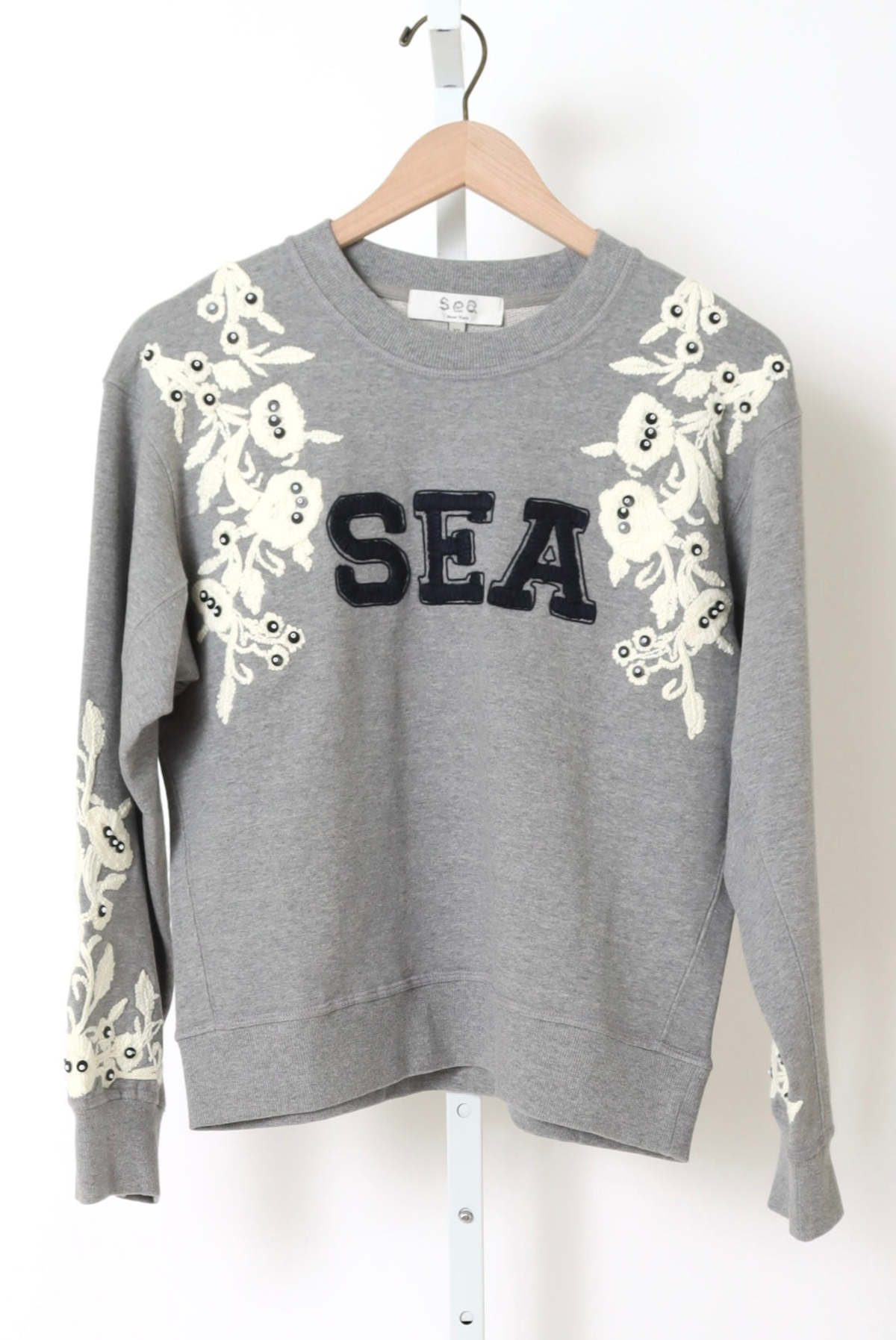 The Georgie Sweatshirt by Sea New York is a gray cotton-polyester blend featuring bold black "SEA" lettering, white floral appliqués, and black beads on the shoulders and sleeves. Displayed on a wooden hanger. True to size; dry clean recommended.
