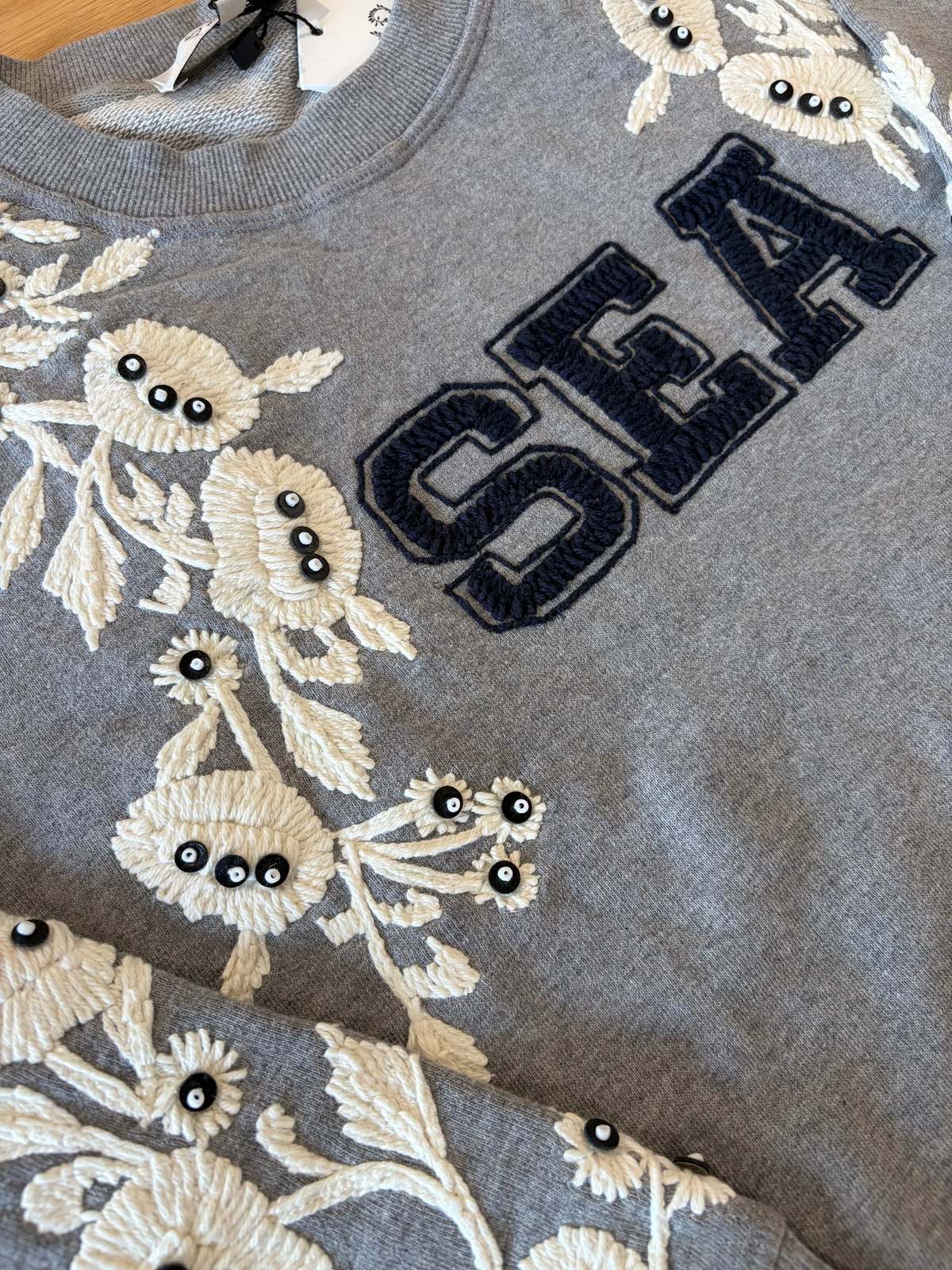 The Georgie Sweatshirt by Sea New York is a gray cotton-poly blend featuring "SEA" in large navy letters, white embroidered flowers, and black bead details on the sleeve and shoulder. True to size—perfect for any casual look.