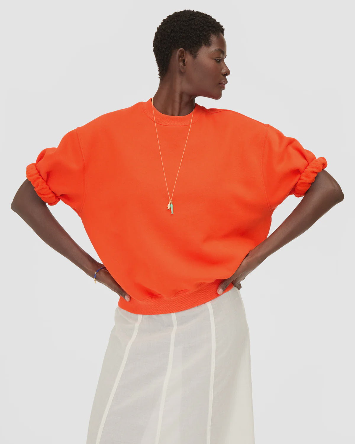 Clare Vivier bright orange oversized short sleeve sweatshirt styled with white skirt, hands on hips.