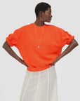 Clare Vivier bright orange oversized short sleeve sweatshirt styled with white skirt, hands on hips.