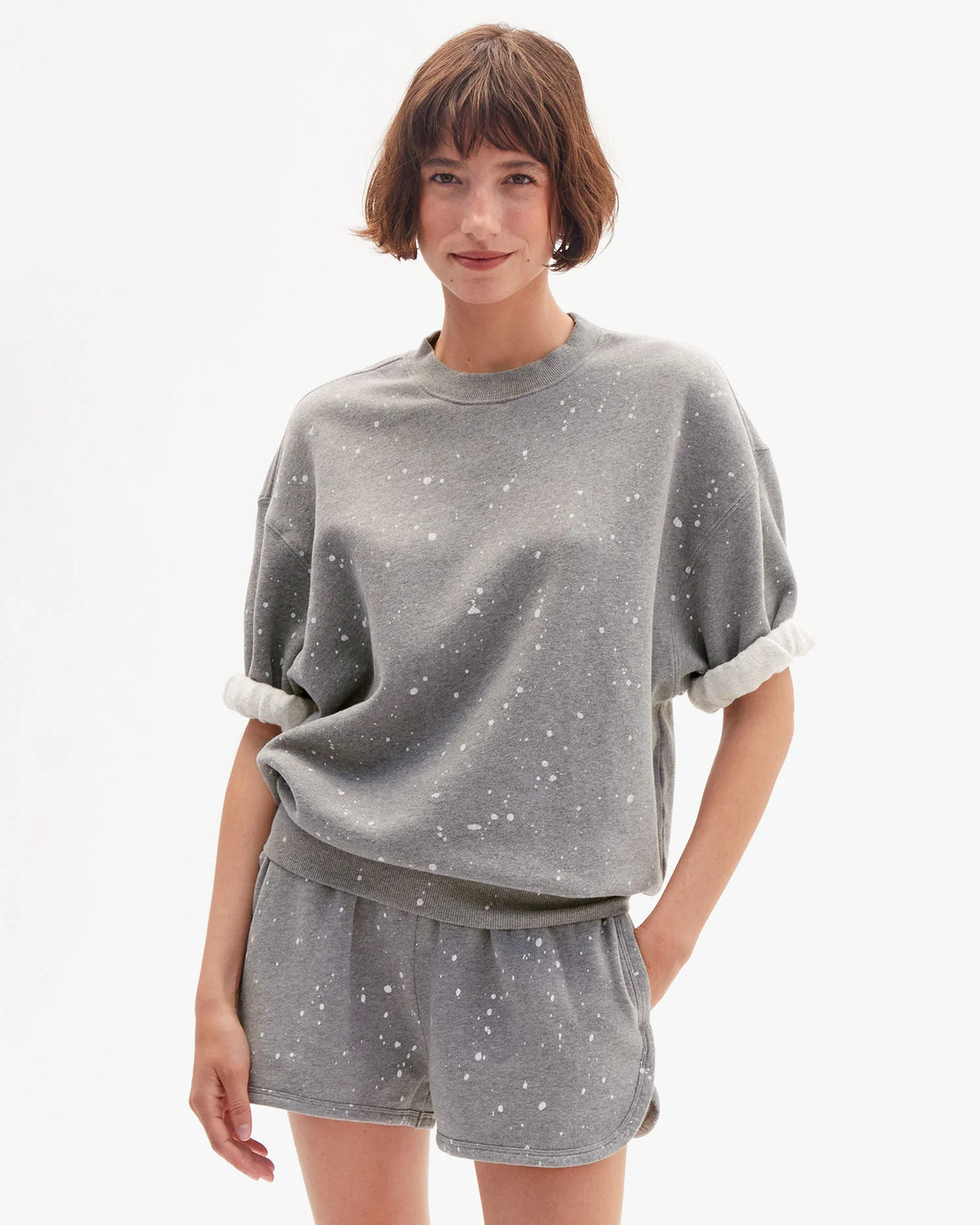 A woman with short brown hair wears the Clare Vivier SS Oversized Sweatshirt in gray cotton fleece with rolled sleeves, paired with matching shorts, standing against a white background and smiling softly.